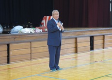 体育館のステージ下で右手にマイクを持つ市長が挨拶を行っている彦成地区の体育祭の様子の写真