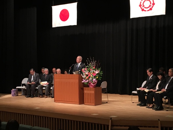 花台に色鮮やかな花が設置されている舞台上で、演台に立って話をする市長と、両脇の椅子に着席している関係者たちの写真
