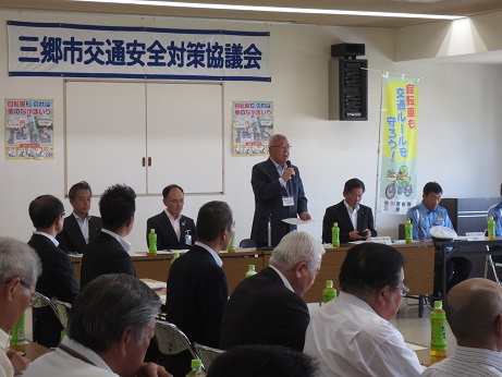 「三郷市交通安全対策協議会」と書かれた横断幕が壁の上部に貼り付けられており、その前に設置された席に立ちあいさつの言葉を述べる三郷市長と、席札とペットボトルのお茶が置かれた席に座り話を聞く出席者の写真