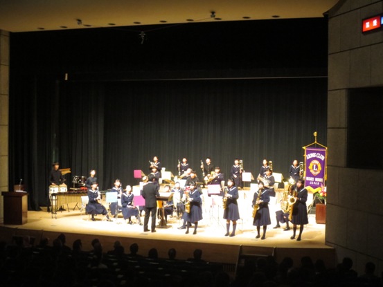 舞台上に吹奏楽の生徒たちが演奏を披露しており、4名の女子生徒が前に出てサックスを吹いている写真