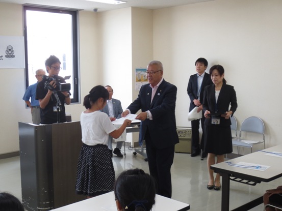 演台の前に立ち小学生の女の子に民生委員・児童委員の委嘱状を手渡す三郷市長と、その様子をビデオカメラで撮影するカメラマンの写真