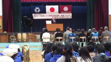 「優れた『地域による学校支援活動』推進にかかる文部科学大臣表彰を祝う会」と書かれた横断幕が飾られたステージの檀上に立つ市長の話をパイプ椅子に座っている方々や床に体育座りをしている子供たちが聞いている後ろ姿の写真