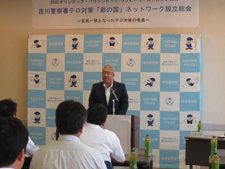 「吉川警察署テロ対策（彩の国）ネットワーク設立総会」と書かれた横断幕が壁に貼り付けられてあり、埼玉警察署のバックボード前の演台に立ちあいさつの言葉を述べる三郷市長と、お茶のペットボトルが置かれた席に座り話を聞く参加者の写真