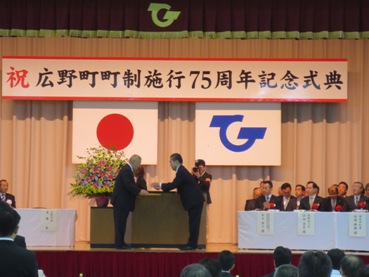 「祝 広野町町制施行75周年記念式典」と書かれた横断幕の演壇の前で遠藤町長と向かい合あった市長が感謝状を頂いている記念式典の様子の写真