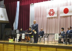 体育館のステージの左右に設置された長机の席に来賓者などが座り、中央に設置されたマイクスタンドの前に立つ市長が挨拶を行っている新三郷地区の体育祭の様子を右斜めから撮影した写真