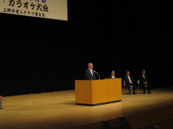 ステージ上には「演芸・カラオケ大会」と書かれた横断幕が吊るされており、演台に立って開会のあいさつの言葉を述べる三郷市長と、ステージ上に置かれたパイプ椅子に座り話を聞く関係者3名の写真