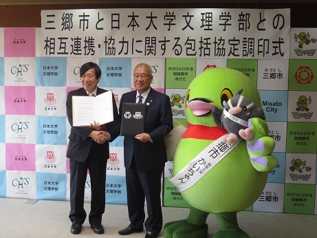 「三郷市と日本大学文理学部との相互連携・協力に関する包括協定調印式」と書かれたポスターが貼りつけられている三郷市と日本大学のバックボードの前で、三郷市長と日本大学の関係者がそれぞれ協定書を手に持ち互いに握手をしながらかいちゃん・つぶちゃんとともに記念撮影をしている写真