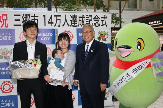 「祝三郷市14万人達成記念式」と書かれたポスターが貼りつけられている三郷市のバックボードの前で、記念品を手に持つ男性と赤ちゃんを抱いた女性が三郷市長とタスキをかけたかいちゃんとともに記念撮影をしている写真