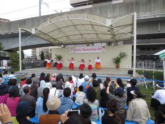 ステージ上に飾られた白地に「Misato Hawai'ian FES」の文字に花のデザインが施されたボードの前で、フラダンスを披露する白いTシャツと赤いパウスカートを着た子どもたちと、芝生の上のベンチに座りダンスの様子を撮影したりして楽しんでいる観客の写真