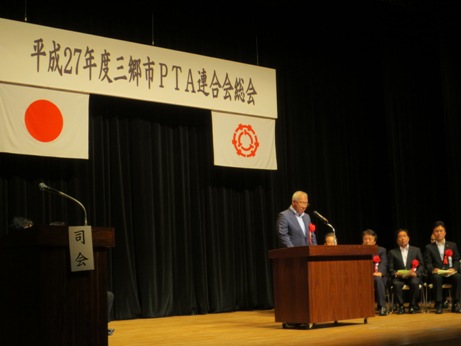 「三郷市PTA連合会総会」と書かれた横断幕が飾られたステージ上の演壇で挨拶を行っている市長の写真