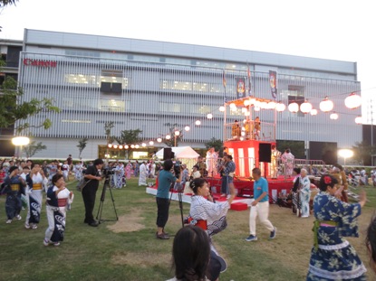 キャノンの会社の建物が隣接する会場の公園中央に櫓が組まれ、周囲を浴衣を着た方々が踊っている様子を2台のテレビカメラで撮影している様子を写した写真