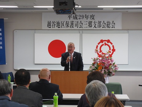 「平成29年度越谷地区保護司会三郷支部会総会」と書かれたポスターが国旗と三郷市旗が飾られたホワイトボードの上に貼ってあり、赤・ピンク・オレンジの花々で彩られた花台と並ぶ演台に立ってあいさつの言葉を述べる三郷市長とペットボトルのお茶が置かれた席に座り話を聞く参加者の写真