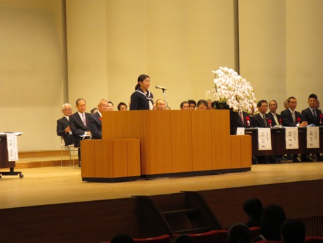 ステージの右側に設置された長机の席に座っている関係者が、檀上で挨拶を行っている森田生徒会長の話を聞いている写真
