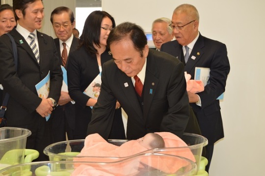 新生児用ベッドに寝かされたピンクのタオルにくるまれている赤ちゃんの人形を抱きかかえる上田清司埼玉県知事と、後ろで説明を聞く三郷市長と関係者の写真