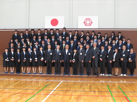 国旗と三郷市旗が飾られている壁の前で、三郷市長と職員が職員証を首にかけた大勢の新入職員とともに並んで記念撮影をしている写真
