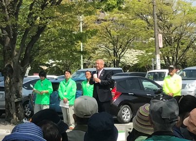 たくさんの木々に囲まれた駐車場のそばであいさつの言葉を述べる三郷市長と、話を聞く緑のジャンパーを着たスタッフと多くの参加者を写した写真