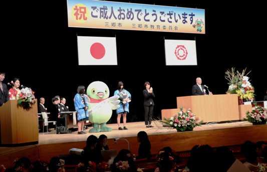 「祝 ご成人おめでとうございます」の看板が設置されている舞台上に、振袖やスーツ姿の司会者や法被姿の関係者、かいちゃんが立っており、演壇に立って話をしている市長の写真