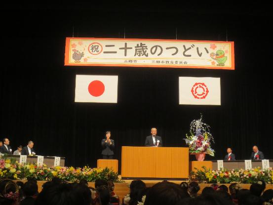 両端にかいちゃん、つぶちゃんが描かれた祝二十歳のつどい横断幕や国旗、市章の下、大きな生花が飾られた壇上で挨拶をする木津市長と手話通訳者の方の写真