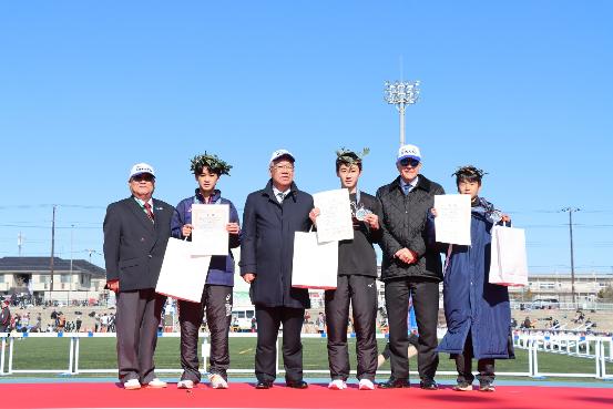 レッドカーペットが敷かれた台の上に三郷市長と駐日ギリシャ大使 、みさとシティハーフマラソン実行委員長、3人の子どもが乗り、メダルと表彰状を持ち一緒に記念撮影をしている写真