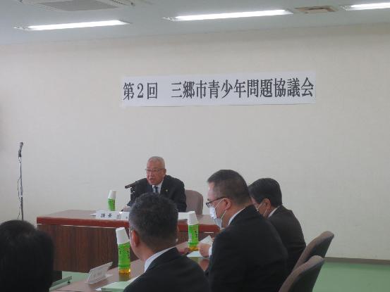 前方に第2回三郷市青少年問題協議会の横断幕が設置された会議室で、自席に座って挨拶をしている三郷市長の写真