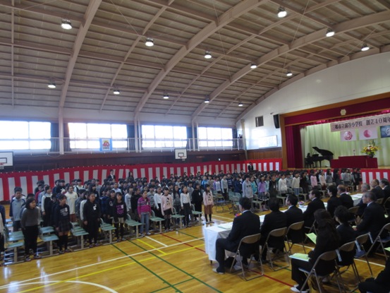 壁に紅白幕が掛けられている体育館で、席を立ち、来賓者席を向いて、ステージのグランドピアノの演奏にあわせ素晴らしい合唱を披露する前谷小学校の在校生265名と、並んだパイプ椅子に座り鑑賞する来賓と関係者の写真