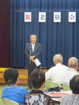 「祝 敬老会」と書かれた文字が飾られた会場で、挨拶を行っている市長と、市長の話を聞いている参加者を後方から写した写真