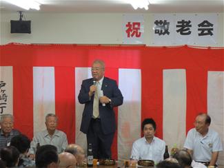 壁に紅白幕が貼られ、マイクを持ち挨拶を行っている市長と、市長の話を聞いている参加者の後ろ姿の写真