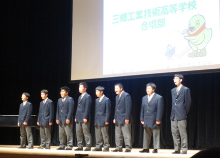 「三郷工業技術高等学校合唱部」の文字とハートをともに持つかいちゃんとつぶちゃんのイラストが映し出されたスクリーンの前で、制服を着て横一列に並び合唱を披露している三郷工業技術高等学校合唱部の男子学生たちの写真