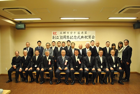 「祝三郷市空手道連盟創立30周年記念式典・祝賀会」と書かれたポスターが貼り付けてある壁の前で、三郷市長と三郷市空手道連盟関係者が並んで記念撮影をしている写真