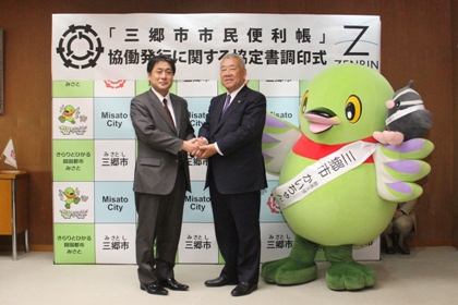 「「三郷市市民便利帳」協働発行に関する協定書調印式」と書かれたバックパネルの前右側にマスコットキャラクターかいちゃんとつぶちゃん、中央に男性と握手をしている市長の写真