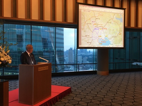 三郷市長が演台に立ち、ガラス張りの壁に設置されたスクリーンに映し出されている三郷市広域図を用いながら三郷市をPRしている日本ギリシャ協会総会の様子の写真