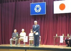 国旗と三郷市旗が飾られた体育館のステージの左右に設置された長机の席に来賓者などが座り、中央に設置されたマイクスタンドの前に立つ市長が挨拶を行っている早稲田地区の体育祭の様子の写真