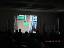 スクリーンに建物の柱に設置された緑色の看板の画像が映し出された様子を見ている参加者の後ろ姿の写真