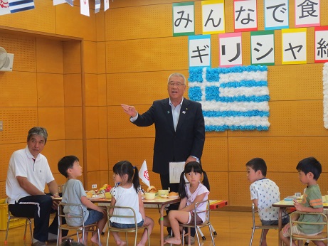 赤・黄色・緑・水色・ピンクの台紙に一文字ずつ貼られた「みんなで食べようギリシャ給食」の文字と、おはながみで作られたギリシャの国旗が壁に飾られている室内で、ギリシャ料理の給食が並ぶ子供用のテーブルに着席した子どもたちと、テーブルの方を向いて立ち、話をする三郷市長の写真
