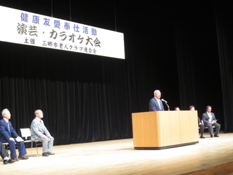「演芸・カラオケ大会」と書かれた横断幕下のステージ左右に設置された椅子に各3名ずつ座り、中央の演壇に立ち挨拶を行っている市長の写真