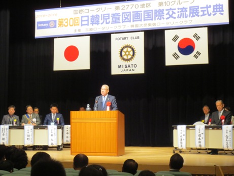 「第30回日韓児童図画国際交流展式典」と書かれた横断幕と日本と韓国の国旗が飾られたステージの演壇で、胸章リボンをつけた市長が挨拶を行っている写真