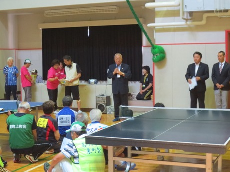 卓球台が設置されている大会の会場の中央に立つ市長の話を床に座って聞いている参加者の後ろ姿の写真