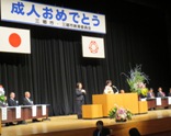 「成人おめでとう」と書かれtた横断幕が飾られたステージの檀上で、振り袖姿の女性が二十歳の主張を行っている様子を左斜めから撮影した写真
