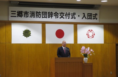 「三郷市消防団辞令交付式・入団式」と書かれた横断幕と国旗と三郷市旗などが壁に飾られた室内の演壇に立つ市長が挨拶を行っている写真