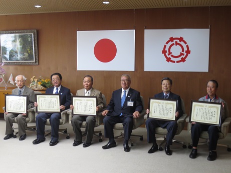国旗と三郷市旗が吊るしてある壁の前で、三郷市長を中心に、感謝状を両手に持った男性5名が椅子に座り記念撮影をしている写真