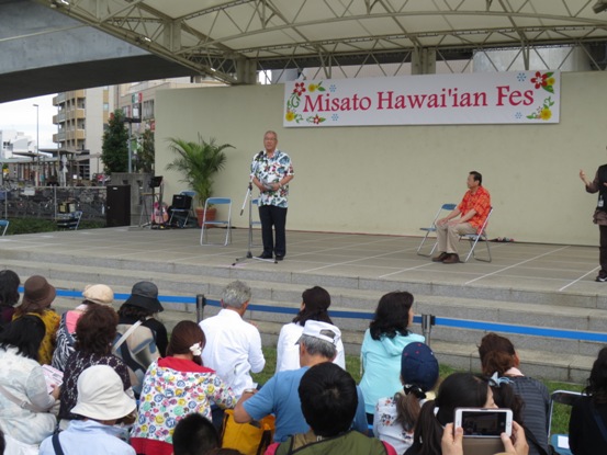 ステージ上に飾られた白地に「Misato Hawai'ian Fes」の文字に花のデザインが施されたボードの前で、式典のあいさつの言葉を述べるアロハシャツを着用した三郷市長と、芝生の上に座り話を聞く参加者の写真