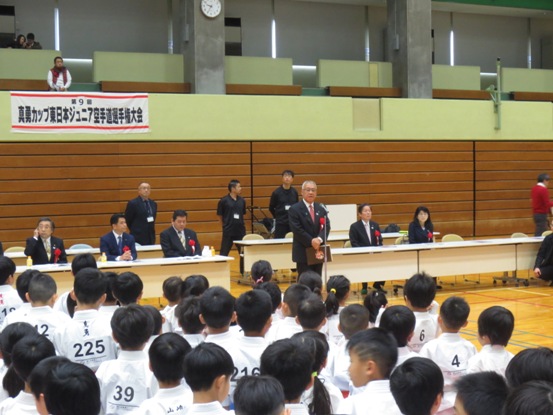 「第9回真勇カップ東日本ジュニア空手道選手権大会」と書かれた横断幕が壁に貼り付けてあり、前に立って激励の言葉を述べる三郷市長と、話を聞くゼッケンが付いた空手着を着用した大勢の子どもたち、壁側に配置された席に座る関係者の写真