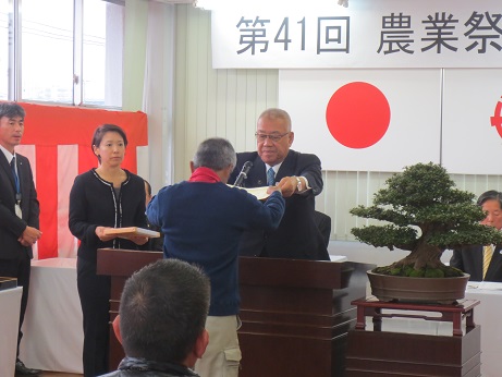 「第41回三郷市農業祭表彰式」と書かれた横断幕と国旗・三郷市旗が貼り付けられている壁の前で、立派な盆栽が飾られている花台と並ぶ演台に立ち表彰状を手渡す三郷市長と受け取る男性、演台の近くに立つ記念品を持った介添人の女性の写真