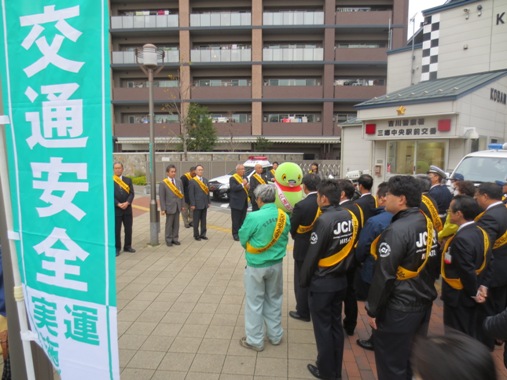 交番の前に黄色のタスキを肩にかけた関係者が集まり、中央に立っている市長が挨拶を行っている様子の写真