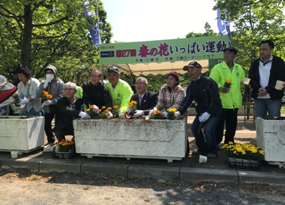 「第27回春の花いっぱい運動」と書かれた横断幕の前で、三郷市長が緑のジャンパーを着たスタッフと参加者とともにオレンジや黄色の花を白いプランターに植え付けている写真
