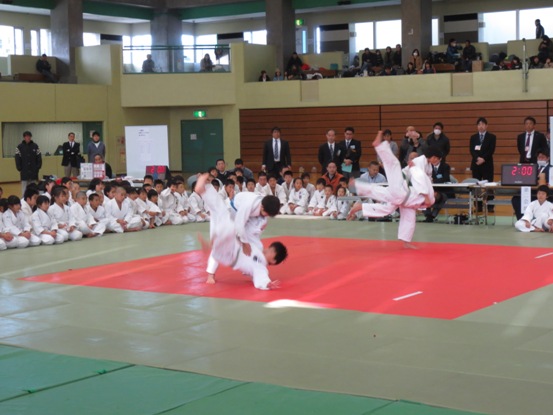 体育館の床に敷かれた赤いシートの上で素晴らしい演武を披露する2組の子どもたち、その様子を少し離れたところに並んで座り見学する柔道着姿の子どもたちと、その後ろに立つ関係者たちの写真