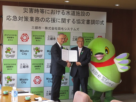 「災害時等における水道施設の応急対策業務の応援に関する協定書調印式」と書かれたポスターが貼られた三郷市のバックボードの前で、三郷市長と株式会社両毛システムズの秋山社長が協定書を片方ずつ持ち合いながら握手をし、かいちゃんとともに記念撮影をしている写真