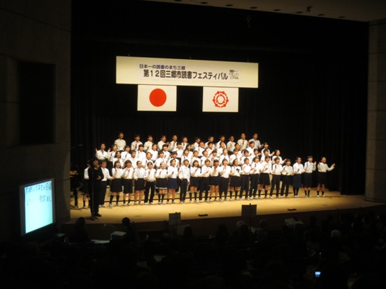 「日本一の読書のまち三郷 第12回三郷市読書フェスティバル」の吊り看板が設置された舞台上で、制服を着た生徒たちが横4列に並んで合唱を披露している様子の写真