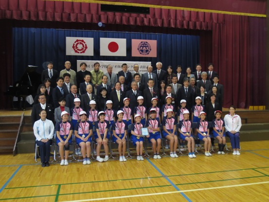「第4回全国小学校ラジオ体操コンクール表彰式」と書かれた横断幕と国旗・三郷市旗・彦成小学校の校章旗が吊り下げられているステージの前で、紅白帽と体操服を着用し金メダルを首に掛けた彦成小学校の子どもたちと三郷市長、関係者が並んで記念撮影をしている写真