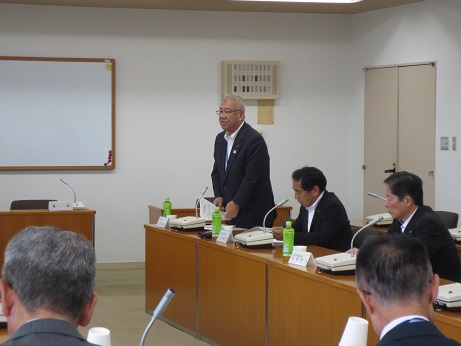 席に立ち資料を両手に持ちながら話をする三郷市長と、席札とマイク、ペットボトルのお茶が置かれている席に座り資料に目を通しながら話を聞く参加者の写真